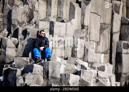 Ein Mann sitzt im Winter auf Basaltsäulen am schwarzen Sandstrand Reynisfjara in der Nähe von Vík í Mýrdal, Südisland Stockfoto