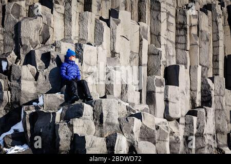 Eine Frau, die im Winter auf Basaltsäulen am schwarzen Sandstrand Reynisfjara in der Nähe von Vík í Mýrdal, Südisland, sitzt Stockfoto