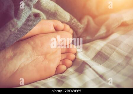 Die Beine eines schlafenden Mannes ragen unter einer karierten Decke hervor und liegen auf einem weichen karierten, von Sulight beleuchteten Laken. Fauler Morgen. Stockfoto