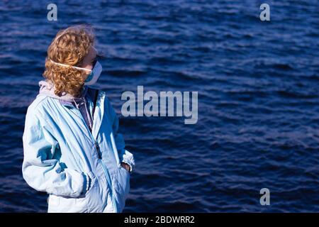 Junges Mädchen mit Händen in Jackentaschen steht auf einem Hintergrund aus blauem Wasser und schaut zur Seite. Frau in einer schützenden Gesichtsmaske vor Viren Stockfoto