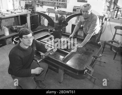 1980s, historisch, zwei männliche Möbelrestauratoren in ihrer kleinen Werkstatt, mit einem antiken Holztisch, kopfüber auf einen anderen Tisch geklemmt, für einfachen Zugang zu den Rahmen und Beine, England, Großbritannien. Stockfoto