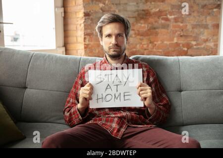 Halten Banner ZU HAUSE bleiben sitzen auf dem Sofa im Zimmer. Kaukasischer Mann zu Hause während der Quarantäne wegen Coronavirus, COVID-19 Ausbreitung. Konzept der Medizin und Gesundheit, Isolation. Stockfoto