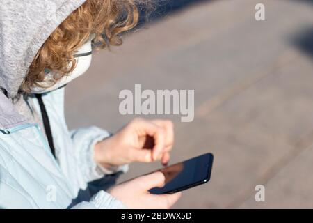 Frau in Kapuze und Jacke benutzt ein Telefon auf der Straße. Frau mit medizinischer Schutzmaske. Das Konzept des technologischen Fortschritts in Stockfoto