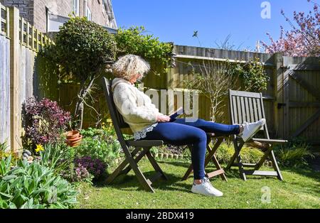 Frau mittleren Alters, die während der COVID-19 UK ein Buch in ihrem Garten liest, während sie sich zu Hause selbst isoliert Stockfoto