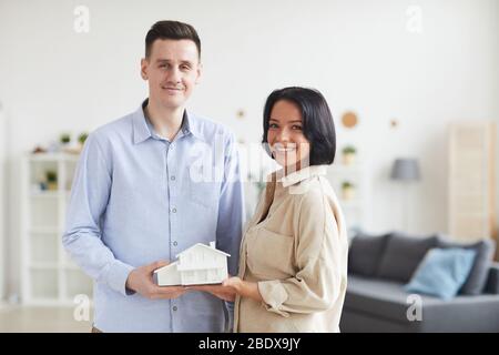 Porträt eines jungen Paares, das das Modell des Hauses in den Händen hält und beim Stehen im Wohnzimmer zu Hause die Kamera anlächelt Stockfoto