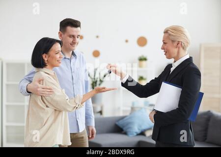 Reife Immobilienmakler geben Schlüssel aus dem neuen Haus zu jungen Paar, während sie im Wohnzimmer zu Hause stehen Stockfoto