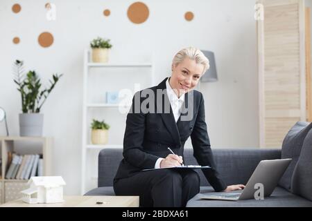 Porträt einer reifen Geschäftsfrau in schwarzem Anzug lächelnd vor der Kamera, während sie auf dem Sofa sitzt, Notizen macht und Laptop-Computer in ihrer Arbeit verwendet Stockfoto