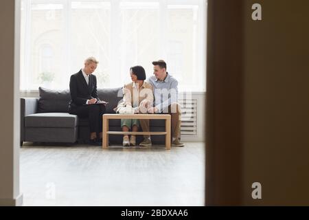 Reife Immobilienmakler in schwarzem Anzug sitzen auf dem Sofa zusammen mit jungen Paar und Unterzeichnung des Vertrags Stockfoto