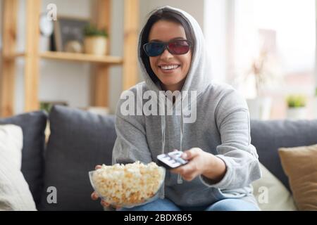 Lächelnde junge Frau in 3D-Brille, die Popcorn isst und zu Hause auf dem Sofa Fernsehen sieht Stockfoto