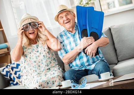 Ein älteres Paar sitzt im Reisebüro und trägt Hüte und eine Schwimmbrille, hält Flossen und sieht fröhlich aus Stockfoto