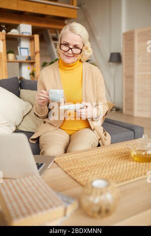 Reife Frau mit blonden Haaren und in Brillen, die Tee trinkt, während sie im Wohnzimmer auf dem Sofa sitzt Stockfoto