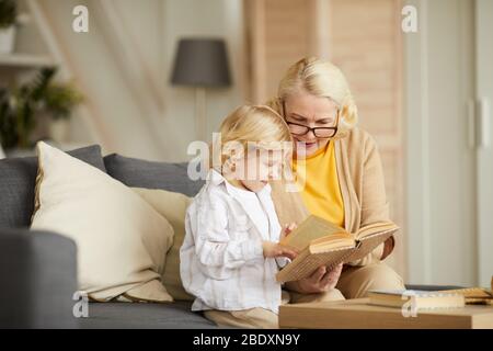 Großmutter trägt Brille und liest ihrem Enkel ein Buch, während sie auf dem Sofa im Wohnzimmer sitzt Stockfoto