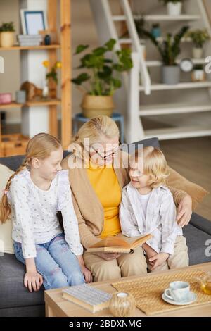 Großmutter liest ihren Enkelkindern ein Buch vor, während sie im Wohnzimmer auf dem Sofa sitzen Stockfoto