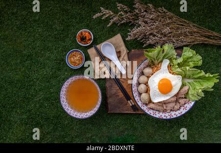 Eiernudeln mit Schweinefleischkugel, Hackfleisch, Spiegelei, Gemüse und Suppen in Schüssel und Essstäbchen mit Löffel und Holz Hintergrund kochen. Thailändische Nudelsuppe Stockfoto