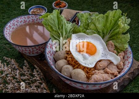 Eiernudeln mit Schweinefleischkugel, Hackfleisch, Spiegelei, Gemüse und Suppen in Schüssel und Essstäbchen mit Löffel und Holz Hintergrund kochen. Thailändische Nudelsuppe Stockfoto
