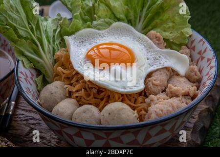 Eiernudeln mit Schweinefleischkugel, Hackfleisch, Spiegelei, Gemüse und Suppen in Schüssel und Essstäbchen mit Löffel und Holz Hintergrund kochen. Thailändische Nudelsuppe Stockfoto