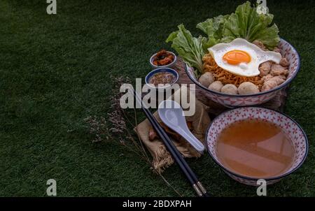 Eiernudeln mit Schweinefleischkugel, Hackfleisch, Spiegelei, Gemüse und Suppen in Schüssel und Essstäbchen mit Löffel und Holz Hintergrund kochen. Thailändische Nudelsuppe Stockfoto