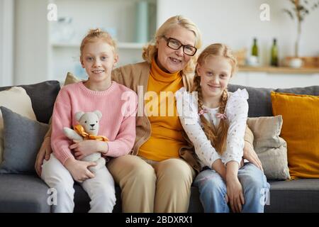 Porträt der glücklichen Großmutter, die auf dem Sofa sitzt und ihre beiden Enkelinnen umarmt und sie lächelnd zu Hause vor der Kamera lächelt Stockfoto