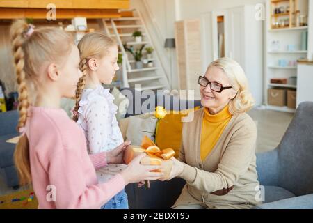 Glückliche Großmutter in Brillen, die von ihren Enkelkindern geschenkt wird, während sie zu Hause im Wohnzimmer sind Stockfoto