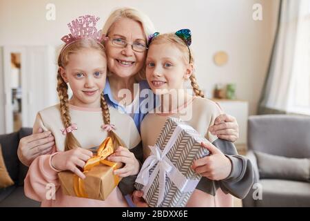 Porträt der Großmutter umarmt ihre beiden Enkelinnen mit Geschenkschachteln und sie lächeln auf Kamera Stockfoto