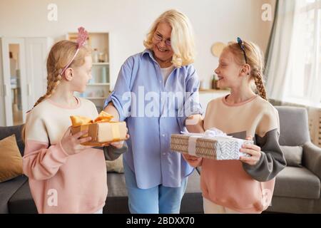 Glückliche Großmutter, die ihren Enkelinnen Geschenkschachteln gibt und ihnen zu ihrem Geburtstag zu Hause gratuliert Stockfoto