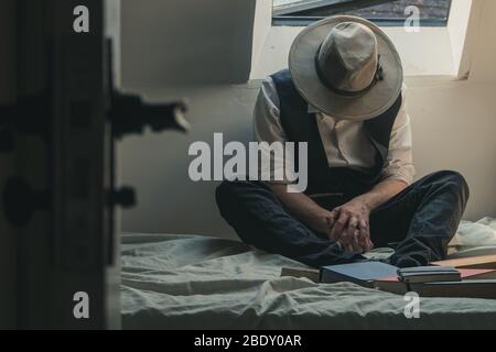 Ein Mann, der mit einem Fedora-Hut und einer Weste auf einem Bett sitzt und Bücher um ihn herum trägt. Stockfoto