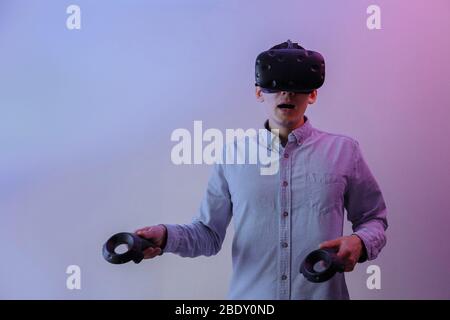 Ein Mann in einer Virtual-Reality-Brille mit Gamepads in den Händen beherrscht die virtuelle Realität. Ein Mann wird von einem Neonlicht beleuchtet. Stockfoto