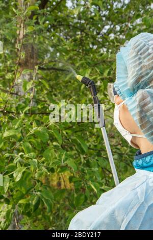 Apfelbaum wird von Chemikalien von Pilzkrankheiten oder Ungeziefer von einer Farmerin in einem Schutzanzug und einer Maske mit einem Drucksprüher in der gesprüht Stockfoto