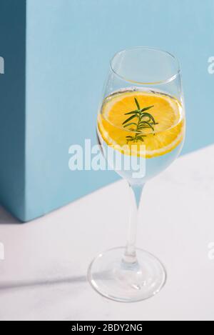 Weinglas mit Sommer hausgemachten kalten Cocktail mit Zitronenscheibe und grünem Zweig auf einem weißen Tisch mit Schatten gegen pastellblaue Wand., Kopierraum. Alco Stockfoto