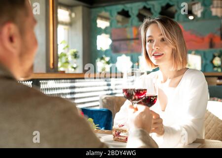 Glücklich junge amorous Frau klirrend Weinglas mit dem von ihrem Freund über servierten Tisch während beide genießen das Mittagessen Stockfoto