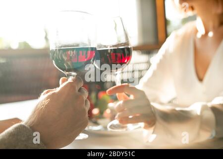 Die Hände zweier junger, amoröser Datteln klatschen mit Gläsern Rotwein, während sie am Tisch im luxuriösen Restaurant sitzen Stockfoto