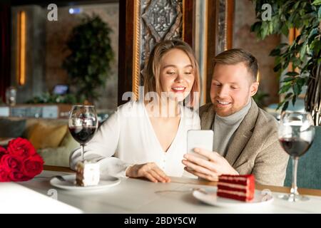 Junge lächelnde Paar etwas in Smartphone beobachten, während sie am Tisch in edlem Restaurant sitzen und Wein und Kuchen Stockfoto