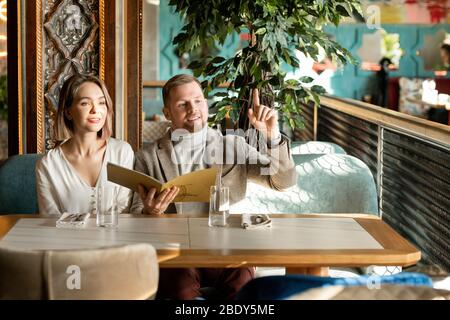 Junger Mann mit erhöhtem Zeigefinger, der darum bittet, dass der Kellner im Restaurant Ordnung bringen kann, während er neben seiner Freundin auf der Couch sitzt Stockfoto