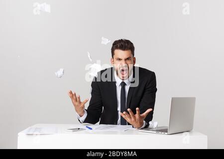 Wütender junger bärtiger Mann arbeitet am Schreibtisch mit Laptop isoliert auf weißem Hintergrund. Schreien reißen Papierdokumente Stockfoto