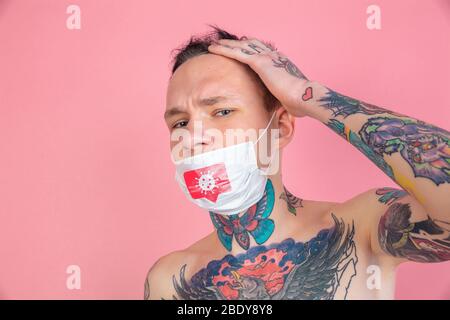 Junger Mann weint auf rosa Hintergrund trägt Gesichtsmaske mit Coronavirus Illustration. Konzept der Verbreitung, Schutz, Prävention, weltweiten Epidemie und Pandemie des Coronavirus. Quarantäne, Isolierung. Stockfoto