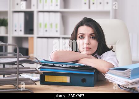 Junge unglückliche Buchhalterin oder Sekretärin, die auf dicken Stapel von Finanzpapieren liegt, während sie vor der Kamera am Schreibtisch sitzt Stockfoto