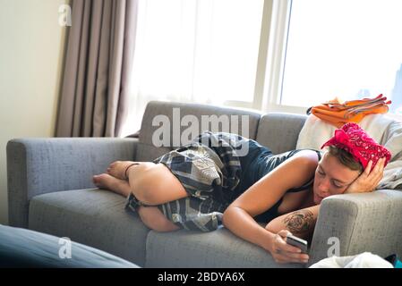 Junge blonde Frau mit Tattoos, die auf einem Sofa ausgestreckt ist, mit ihrem Handy in der Hand und in Hindu-Kleidung gekleidet Stockfoto