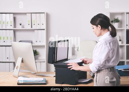 Hübsche junge elegante Sekretärin, die bei xerox Maschine steht und Kopien von Finanzpapieren anfertigen, während sie ihre Arbeit im Büro macht Stockfoto