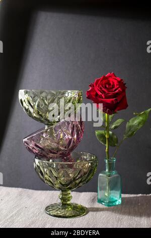 Drei Vasen für creme aus dunkelgrünem und lila Glas stehen übereinander auf dem schwarzen Hintergrund mit Schatten. Dunkelrote Rose im Flascheninste Stockfoto