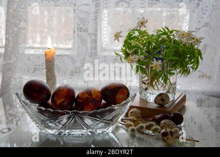 Ein Blumenstrauß aus Frühlingsblumen in einem Glas, bemalte Ostereier an einer gestrickten Serviette vor dem Hintergrund weißer Spitzen-Vorhänge. Stockfoto