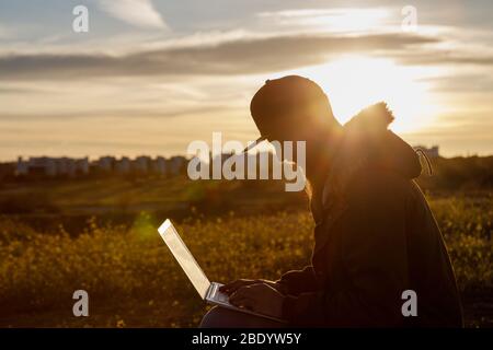 Silhouette eines jungen Mannes mit Bart, der mit seinem Laptop bei Sonnenuntergang in der Natur arbeitet Stockfoto