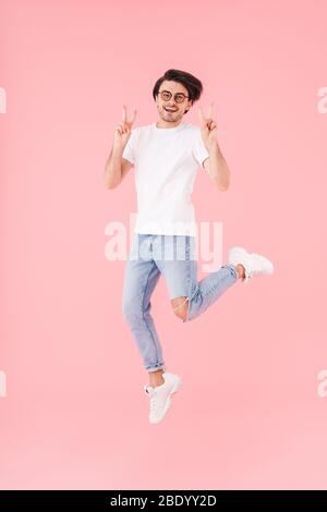 Bild von gutaussehenden glücklichen Mann trägt Brille springen und Gestik Friedenszeichen isoliert über rosa Hintergrund Stockfoto