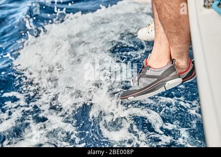 Die Füße des Matrosen hängen von einem Boot Bord über azurblauem Wasser, Schuhe für Yacht, Spritzer Wasser, Sonnenflecken des Lichts auf dem Wasser, Segeln, sonnig Stockfoto