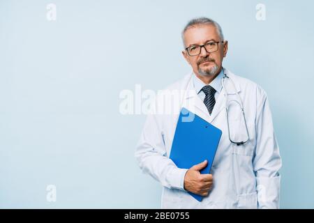 Älterer bärtiger Arzt in weißer Uniform mit Stethoskop und blauer Mappe. Arzt auf dem Hintergrund eines medizinischen Forschungslabors. Stockfoto