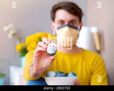 Kaukasischer Mann hält ein osterei mit einer Gesichtsmaske während der osterzeit, Coronavirus Ausbruch Zeit, mit gelben sweather und Blumen im Hintergrund. Stockfoto