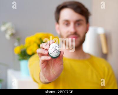 Kaukasischer Mann hält ein osterei mit einer Gesichtsmaske während der osterzeit, Coronavirus Ausbruch Zeit, mit gelben sweather und Blumen im Hintergrund. Stockfoto