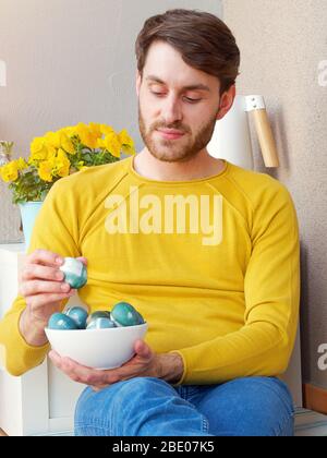 Kaukasischer Mann hält eine blaue ostereier in einer Schüssel während der osterzeit, mit natürlichen Rotkohl Färbung gefärbt, trägt eine gelbe sweather. Stockfoto