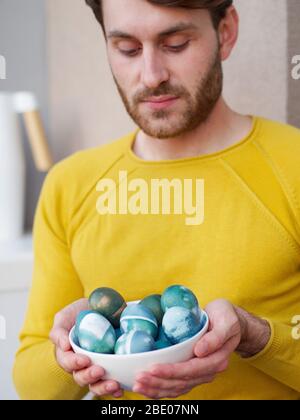 Kaukasischer Mann hält eine blaue ostereier in einer Schüssel während der osterzeit, mit natürlichen Rotkohl Färbung gefärbt, trägt eine gelbe sweather. Stockfoto