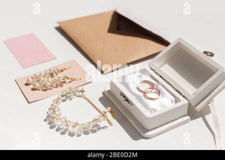 Hochzeit Accessoires Hintergrund. Goldene Eheringe und feminine Accessoires. Goldenes Lametta, Schachtel, Ohrringe, Umschlag, Ringe, Barrette auf weißem Backgr Stockfoto
