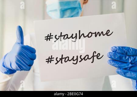 Bleib zu Hause, sichere Hashtags. Kaukasisches Mädchen trägt medizinische Maske und blaue Handschuhe mit weißem Papier mit "Tag-Home-Kampagne" Zeichen. Coronavirus aus Stockfoto